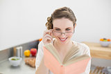 Cheerful woman reading a book while wearing glasses and sitting in her kitchen