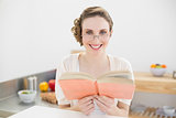 Cheerful brunette woman wearing glasses for reading a book while sitting in her kitchen