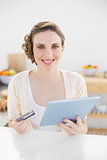 Gorgeous woman using her tablet for home-shopping sitting in her kitchen
