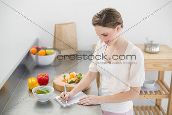 Happy beautiful woman standing in her kitchen writing on a notebook