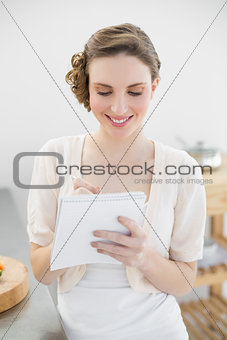 Content lovely woman standing in her kitchen writing a shopping list