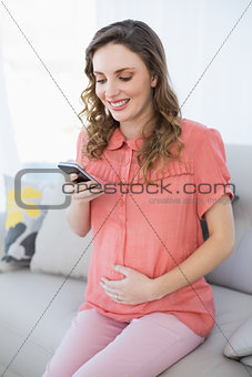 Cheerful pregnant woman using her smartphone sitting on a couch in the living room