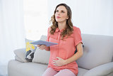 Thinking pregnant woman holding her tablet while sitting on a couch in the living room