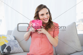 Smiling pregnant woman shaking pink piggy bank sitting on couch