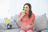 Lovely pregnant woman eating green apple sitting on couch in living room