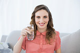 Pretty pregnant woman showing glass of water sitting on couch