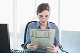 Concentrated young businesswoman reading newspaper sitting at her desk