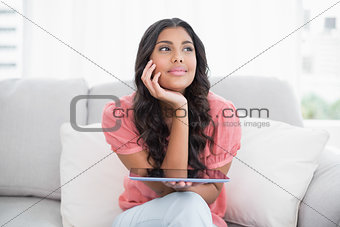 Pensive cute brunette sitting on couch holding tablet