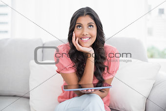 Day dreaming cute brunette sitting on couch holding tablet