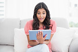 Excited cute brunette sitting on couch using tablet