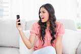 Angry cute brunette sitting on couch holding smartphone