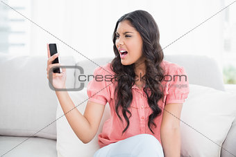 Angry cute brunette sitting on couch holding smartphone