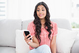 Surprised cute brunette sitting on couch holding smartphone