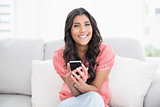 Cheerful cute brunette sitting on couch holding smartphone