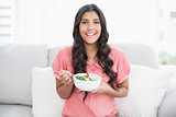Happy cute brunette sitting on couch holding salad bowl