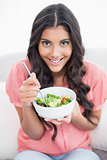 Cheerful cute brunette sitting on couch holding salad bowl