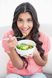 Smiling cute brunette sitting on couch holding salad bowl