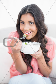 Gleeful cute brunette sitting on couch holding salad bowl