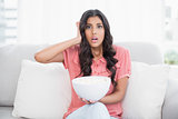 Shocked cute brunette sitting on couch holding popcorn bowl