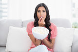 Frowning cute brunette sitting on couch holding popcorn bowl