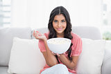 Content cute brunette sitting on couch holding popcorn bowl