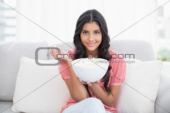 Content cute brunette sitting on couch holding popcorn bowl
