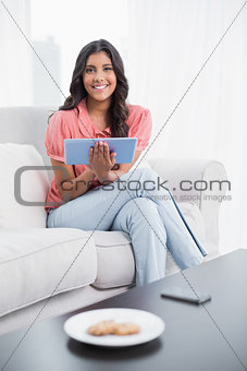 Smiling cute brunette sitting on couch holding tablet