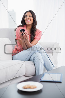 Happy cute brunette sitting on couch using smartphone