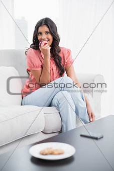 Happy cute brunette sitting on couch eating red apple