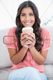 Cheerful cute brunette sitting on couch holding disposable cup