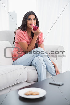 Happy cute brunette sitting on couch holding red apple