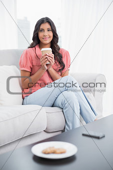 Pleased cute brunette sitting on couch holding disposable cup