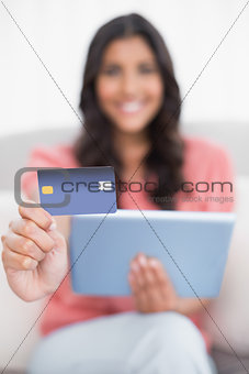 Happy cute brunette sitting on couch showing credit card holding tablet