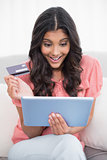 Excited cute brunette sitting on couch holding credit card and tablet