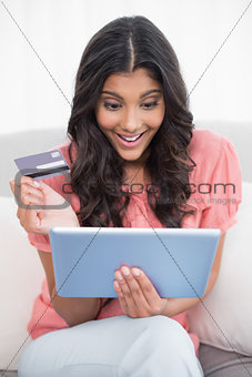 Excited cute brunette sitting on couch holding credit card and tablet