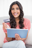 Cheerful cute brunette sitting on couch holding credit card and tablet