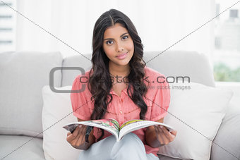 Happy cute brunette sitting on couch holding magazine