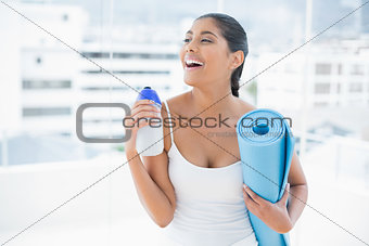 Laughing toned brunette holding sports bottle and exercise mat
