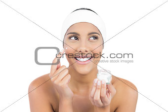Smiling nude brunette using moisturizer looking away