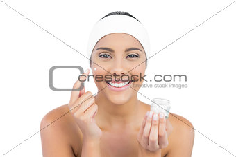 Smiling nude brunette using moisturizer looking at camera