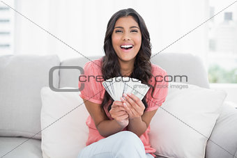Gleeful cute brunette sitting on couch holding money
