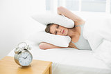 Annoyed young woman holding her ears closed by using pillows while lying in her bed