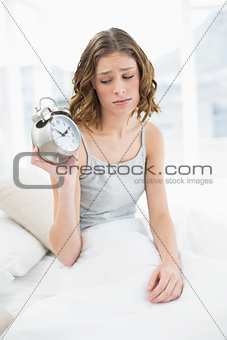 Irritated young woman holding an alarm clock sitting on her bed