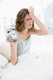 Annoyed young woman holding an alarm clock sitting on her bed