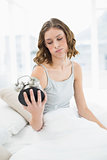Upset woman holding an alarm clock looking at it sitting on her bed