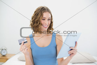 Gorgeous woman holding her tablet and debit card sitting on her bed