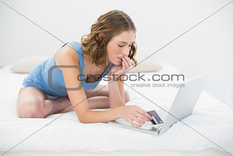 Thinking cute woman sitting on bed while using her notebook for home shopping