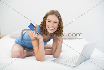 Cheerful beautiful woman showing her credit card while sitting on her bed next to her laptop