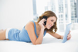 Sweet woman lying smiling on her bed holding her tablet while phoning