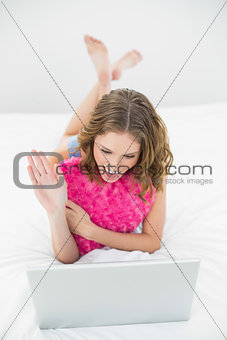 Waving lovely woman using her notebook cuddling with a pillow